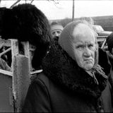 Ältere Person guckt die Betrachtenden direkt an. Links neben ihr auf einem Tor sitzt eine schwarze Katze