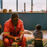 Person sitzt in roter Arbeiterkleidung auf einem Bett. neben ihm sitzt ein Junge, der ihm den Rücken zugekehrt hat