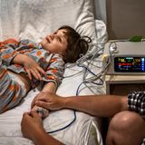 Junges Mädchen liegt auf einem Krankenhausbett und guckt an die Zimmerdecke, während erwachsene person ihre Hand hält