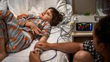 Junges Mädchen liegt auf einem Krankenhausbett und guckt an die Zimmerdecke, während erwachsene person ihre Hand hält
