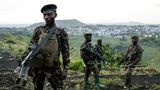 Soldaten stehen in Millitäruniform mit Gewehren auf einem Hügel. Im Hintergrund sieht man eine Stadt