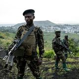 Soldaten stehen in Millitäruniform mit Gewehren auf einem Hügel. Im Hintergrund sieht man eine Stadt