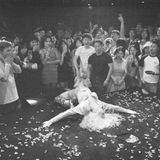 Person liegt auf dem Boden einer Bühne mit ausgebreiteten Armen auf dem Rücken. Um sie herum steht eine Gruppe, die jubelt