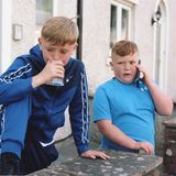 Zwei Jungen stehen vor einem Haus. Einer pustet in einen Platikbecher. Einer hält ein Smartphone an sein Ohr