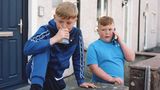 Zwei Jungen stehen vor einem Haus. Einer pustet in einen Platikbecher. Einer hält ein Smartphone an sein Ohr