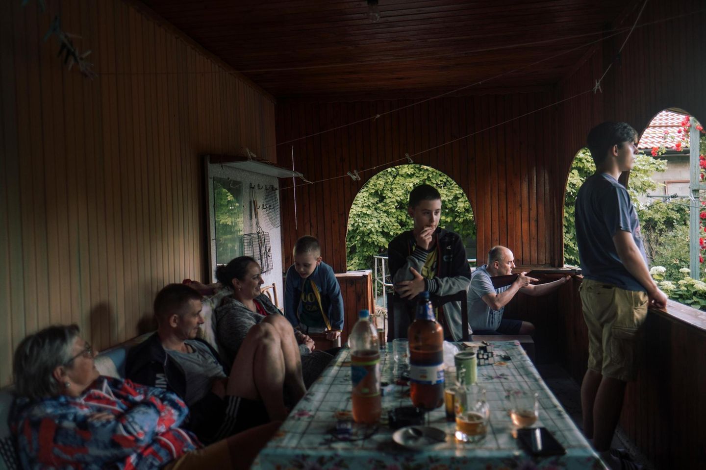 Nostalgie Eine Gruppe Menschen sitzen um einen Tisch herum auf einer Veranda. Eine Person im Hintergrund hält ein Gewehr bereit
