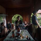 Eine Gruppe Menschen sitzen um einen Tisch herum auf einer Veranda. Eine Person im Hintergrund hält ein Gewehr bereit