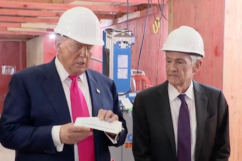 Trump und Powell stehen auf einer Baustelle nebeneinander, beide tragen einen weißen Schutzhelm und Trump hält einen Zettel.