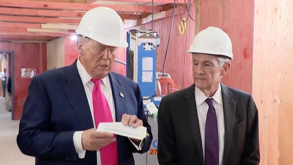 Trump und Powell stehen auf einer Baustelle nebeneinander, beide tragen einen weißen Schutzhelm und Trump hält einen Zettel.