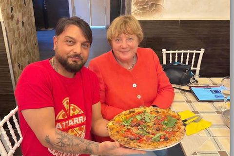 Merkel sitzt in einem roten Obertreil neben einem Pizzeria Mitarbeiter, der eine Pizza hält.