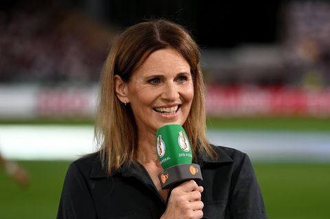Portrait der Fersehmoderatorin Katrin Müller-Hohenstein. Sie steht auf einem Fußballfeld und hält ein Mikrofon in der Hand.