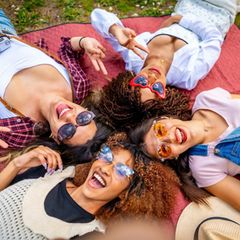Vier Freundinnen liegen auf einer Decke in einem Park, machen ein Selfie und gestikulieren Friedenszeichen