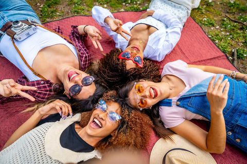 Vier Freundinnen liegen auf einer Decke in einem Park, machen ein Selfie und gestikulieren Friedenszeichen