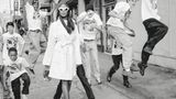 Naomi Campbell stolziert in weißem Mantel und mit weißer Sonnenbrille durch eine Gruppe von Jungen