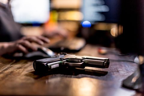 Eine Pistole auf einem Schreibtisch, dahinter unscharf ein Mann am Computer