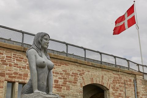 Granitstatue einer Meerjungfrau in Kopenhagen mit großen Brüsten, im Hintergrund eine Dänemark-Flagge