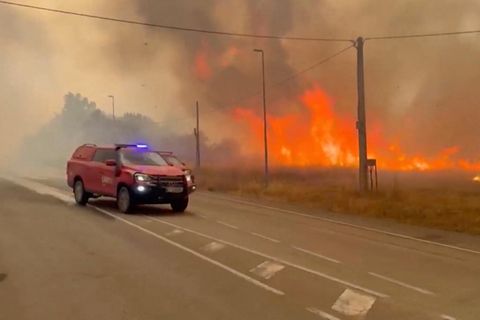 Ein roter Rettungswagen fährt auf einer Straße, im Hintergrung ist ein riesiges Feuer.