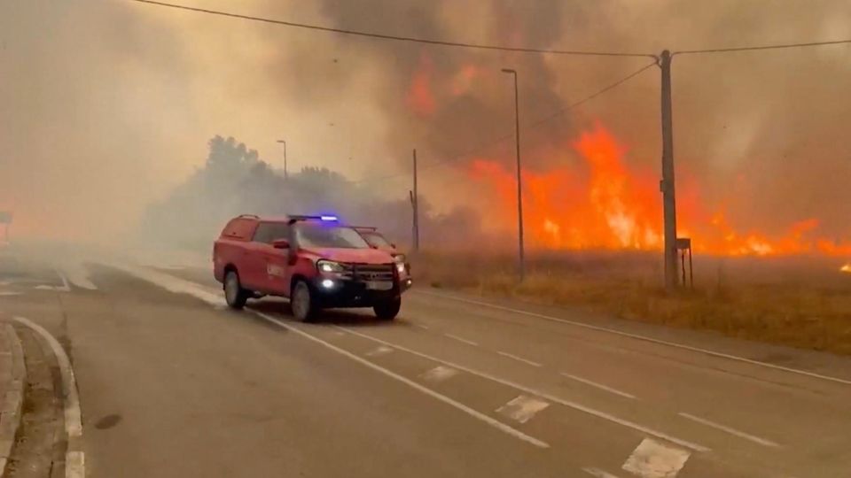 Ein roter Rettungswagen fährt auf einer Straße, im Hintergrung ist ein riesiges Feuer.