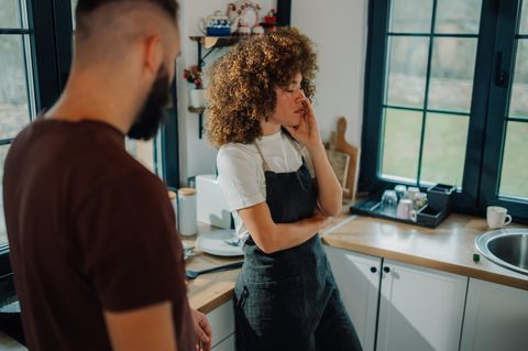 Symbolfoto für eine Affäre: Eine Frau steht neben ihrem Mann in der Küche und hält den Kopf in der Hand