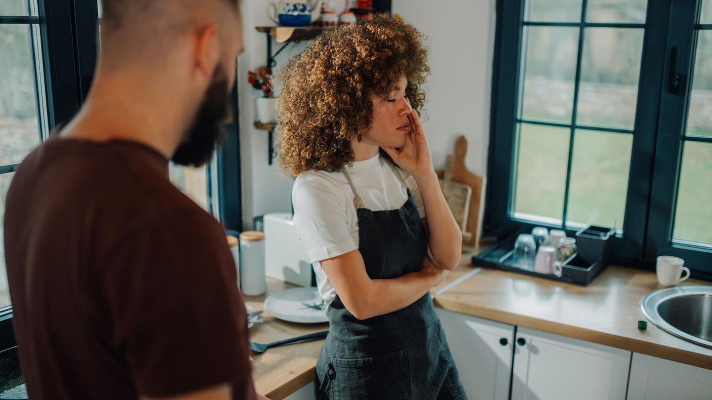 Symbolfoto für eine Affäre: Eine Frau steht neben ihrem Mann in der Küche und hält den Kopf in der Hand