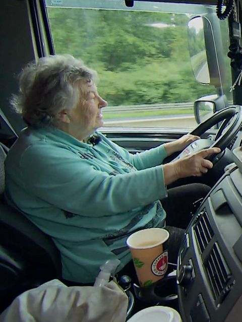 Oma Rosi, gekleidet in einem Pullover sitzt am Lenkrad ihres LKWs und steuert. Vorne hat sie viel Deko stehen.