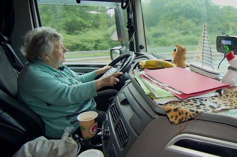 Oma Rosi, gekleidet in einem Pullover sitzt am Lenkrad ihres LKWs und steuert. Vorne hat sie viel Deko stehen.