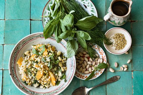 Couscous-Salat mit Aprikosen, daneben ein Teller mit Pistazienkernen und einer mit Basilikum und Dill