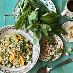 Couscous-Salat mit Aprikosen, daneben ein Teller mit Pistazienkernen und einer mit Basilikum und Dill