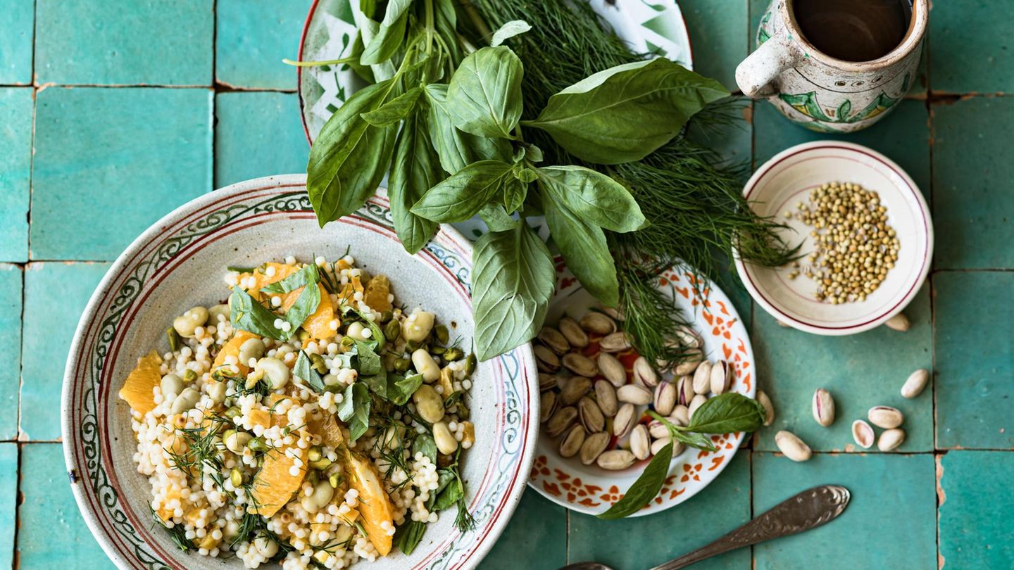 Couscous-Salat mit Aprikosen, daneben ein Teller mit Pistazienkernen und einer mit Basilikum und Dill