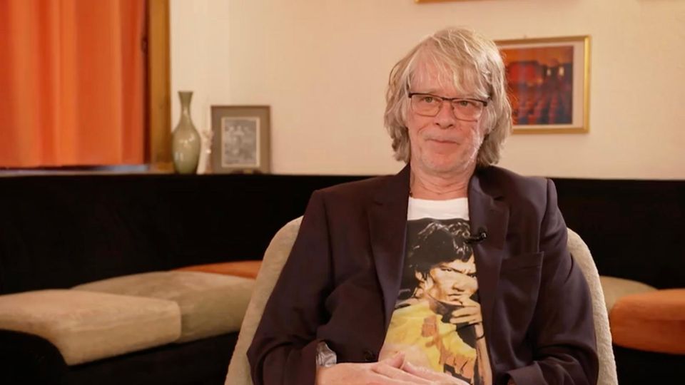 Helge Schneider sitzt mit gefalteten Armen auf einem Stuhl.