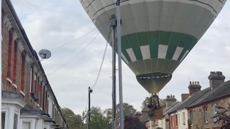 Ein riesiger Heißluftballon schwebt ca 10 Meter über einer Straße mit Wohnhäusern und Autos, Anwohner rennen drauf zu.