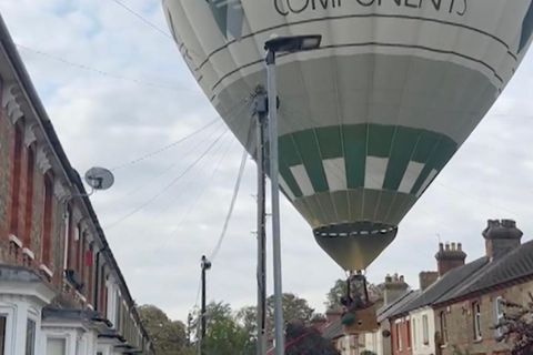 Ein riesiger Heißluftballon schwebt ca 10 Meter über einer Straße mit Wohnhäusern und Autos, Anwohner rennen drauf zu.