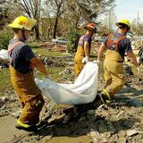 Drei Feuerwehrmänner schleppen einen weißen Leichensack aus einer überschwemmten Nachbarschaft. Mehr als 1300 Menschen verlieren durch Katrina ihr Leben