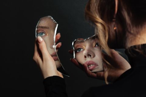 Eine Frau spiegelt ihr Gesicht in Spiegelscherben (Symbolbild Körperbild).