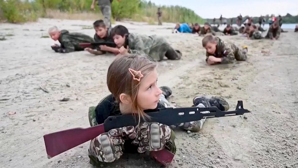 Russische Ferienlager: Kinder trainieren Patriotismus, Patronen und Propaganda Kinder robben in Tarnkleidung und mit Maschinengwehrattrappen über den Sandboden.