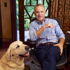 Dan Brown sitzt an einem Holzschreibtisch und lächelt in die Kamera, neben ihm hechelt sein Golden Retriever