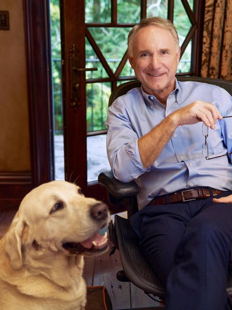 Dan Brown sitzt an einem Holzschreibtisch und lächelt in die Kamera, neben ihm hechelt sein Golden Retriever