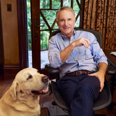 Dan Brown sitzt an einem Holzschreibtisch und lächelt in die Kamera, neben ihm hechelt sein Golden Retriever