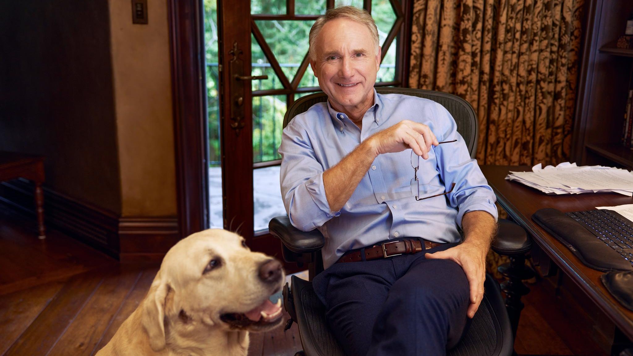 Dan Brown sitzt an einem Holzschreibtisch und lächelt in die Kamera, neben ihm hechelt sein Golden Retriever