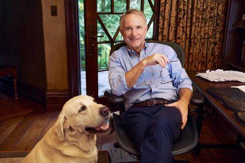 Dan Brown sitzt an einem Holzschreibtisch und lächelt in die Kamera, neben ihm hechelt sein Golden Retriever