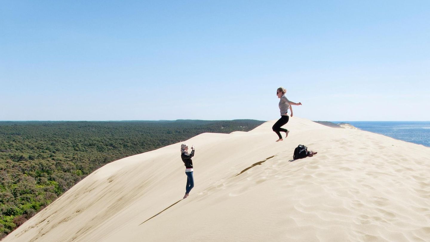 Südfrankreich: Zwei Menschen haben die Wanderdüne Dune du Pilat erklommen und machen Fotos, eine Person springt in die Luft