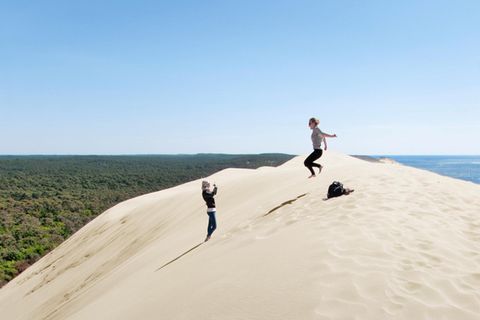 Südfrankreich: Zwei Menschen haben die Wanderdüne Dune du Pilat erklommen und machen Fotos, eine Person springt in die Luft