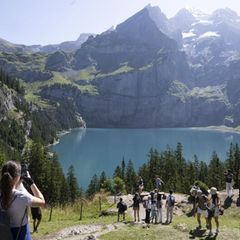 Auswanderziel: Wiese, Blauer Himmel, schneebedeckte Gipfellandschaft in der Schweiz, Wanderer vor Bergsee