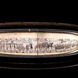 dunkle Ausstellungshalle, mit einem knapp 70 Meter langen bestickten Stoffstreifen in einer beleuchteten Vitrine