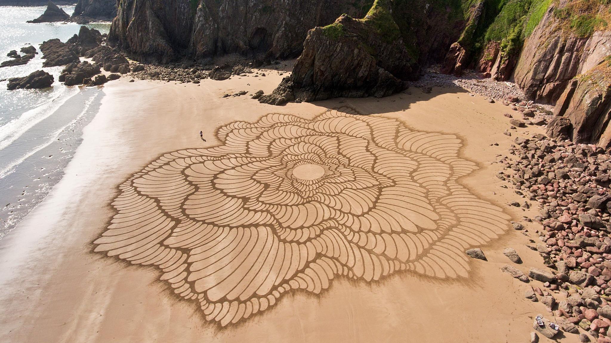"Fluctuo" heißt dieses Werk des Künstlers Jon Foreman, er zeichnete es 2023 am Strand Lindsway Bay in Wales mit einem Laubrechen in den Sand – freihand, bis auf den Kreis in der Mitte