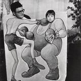 Zwei Personen halten ihre Köpfe durch ein bemaltes Schild auf dem zwei sich boxende Personen zu sehen sind