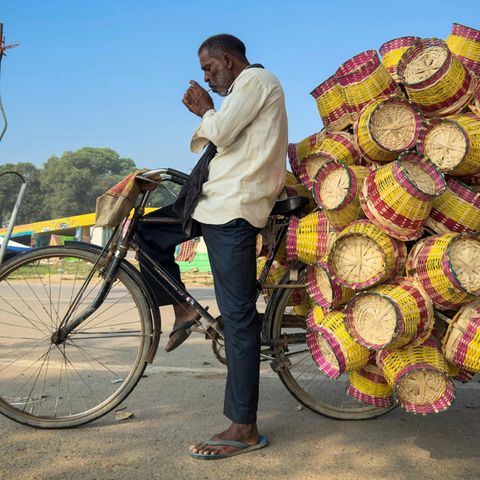 Prayagraj, Indien. Dieser Verkäufer transportiert auf seinem Fahrrad eine ganze Ladung bunter Bambuskörbe durch die Stadt. Doch bevor er weiter Körbe verteilt, gönnt er sich eine Zigarette