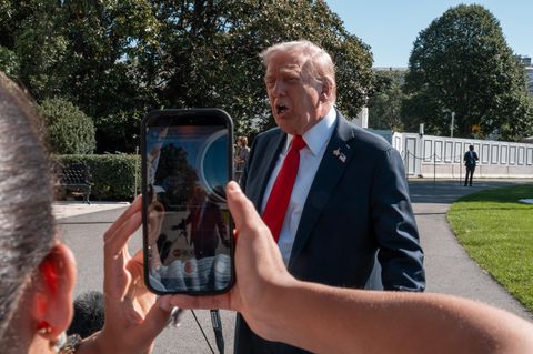 Donald Trump spricht vor der Presse wöhrend eine Frau ihn mit ihrem Handy filmt