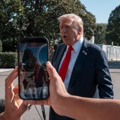 Donald Trump spricht vor der Presse wöhrend eine Frau ihn mit ihrem Handy filmt