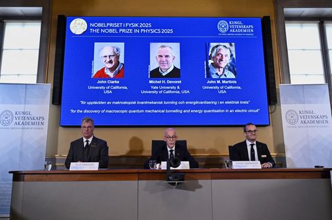 Drei weiße, ältere Männer in Anzügen verkünden die Empfänger des Nobelpreises für Physik 2025